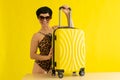 A woman in a solid leopard swimsuit and sunglasses holds a suitcase on a yellow background. A smiling brunette in a Royalty Free Stock Photo