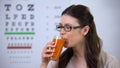 Woman in spectacles drinking fresh carrot juice, eyechart on background, vision