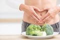 Woman with sports figure on her belly shows heart shape. Fresh broccoli in plate on kitchen table