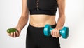 A woman in sportswear holds a dumbbell and avocado in her hands - a healthy diet and sports