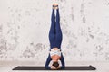 Woman in sportswear practicing yoga performs inverted asana, headstand, shirshasana exercise on mat near grunge wall