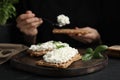 Woman spreading cottage cheese onto crispy cracker at black table, focus on board with sandwiches