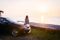 Woman stand by the black luxury car and looks to panoramic view of calm sea and setting sun Royalty Free Stock Photo