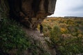 Woman standing by lighted exit in cave, daytime. Atmospheric snapshot in natural rock formations with autumn forest and dramatic