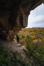 Woman standing by lighted exit in cave, daytime. Atmospheric snapshot in natural rock formations with autumn forest and dramatic