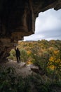 Woman standing by lighted exit in cave, daytime. Atmospheric snapshot in natural rock formations with autumn forest and dramatic