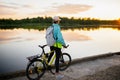 Woman is standing with mountain bike in cross country road at sunset in summer. Looking direct. Colorful lake landscape