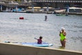 A woman standing in the ocean water using a cell phone with a child on a paddle board and people rowing kayaks in Long Beach