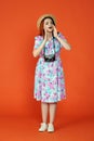 Woman standing with old film camera, looking away and calling for someone, hands over mouth, isolated on orange Royalty Free Stock Photo