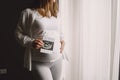 Pregnant woman stands by the window and looks at photo