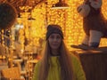 A woman stands in front of a cafe lit by bright evening garlands near a toy decorative lamb. Soft focus