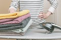 Woman stands by an ironing board with an iron and is holding a stack of ironed, clean terry towels