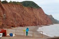 A woman stands by the railings on the east beach at Sidmouth and looks at the red Jurassic cliffs. The cliffs suffer much erosion