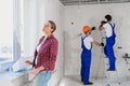 The woman stands at the window and examines the layout of the apartment, builders are working in the background