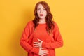 Woman with stomachache, having food poisoning, posing isolated over yellow background, looks at camera, lady has red wavy hair Royalty Free Stock Photo