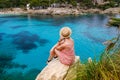 A woman in straw hat sitting on a cliff, looking towards Cala Gat beach in Mallorca