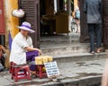 Woman street vendor sits in stool outside entrance to a building selling cakes on a tray