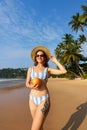 Woman in striped bikini holds fresh coconut on sunny beach. Solo traveler enjoys tropical seaside, healthy drink. Palm