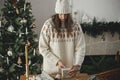 Woman in stylish sweater and hat wrapping christmas gift in paper on wooden table with festive decorations in decorated Royalty Free Stock Photo