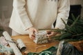 Woman in stylish sweater wrapping christmas gift in paper on wooden table with festive decorations in decorated scandinavian room Royalty Free Stock Photo