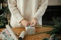 Woman in stylish sweater wrapping christmas gift in paper on wooden table with festive decorations in decorated scandinavian room Royalty Free Stock Photo