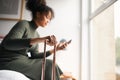 Woman with suitcase checking out of boutique hotel sitting on bed waiting for taxi ordered on mobile phone app Royalty Free Stock Photo