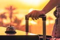 Woman with suitcase ringing hotel service bell with sea and palm tree view on sunset. Travel concept. 24-hour beach hotel front