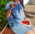 A woman in a summer denim dress is sitting on a park bench, and eating fresh ripe strawberries from a plastic bag bought Royalty Free Stock Photo