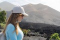 woman with sunglasses and cap looks at the ground in front of volcano