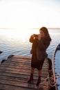 Woman at sunset taking a photo with her mobile on a jetty of a lake with the sun behind