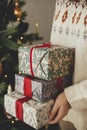 Woman in sweater holding stylish christmas gifts with red ribbon at modern decorated vintage tree in scandinavian room. Merry Royalty Free Stock Photo