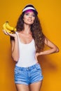 Woman in swimsuit and blue shorts holding banana and posing isolated over yellow background Royalty Free Stock Photo