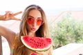 Woman in a swimsuit and red sunglasses with a watermelon shows a peace sign on a background of tropical hotels and the beach Royalty Free Stock Photo