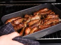 Woman taking black baking tray with baked pork ribs out of an oven