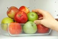 A woman taking a green apple out of a pile of apples that are in a glass tray that is in a kitchen cupboard
