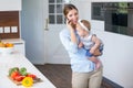 Woman talking on cellphone carrying daughter by kitchen counter Royalty Free Stock Photo