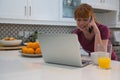 Woman talking on mobile phone while using laptop in kitchen Royalty Free Stock Photo