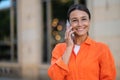 Woman talking by phone wearing orange shirt standing outdoors near building. Royalty Free Stock Photo