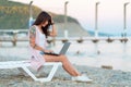 A woman with a tattoo on her arm sits on a chaise longue and works on a laptop. Side view. The concept of remote work and