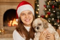 Woman with toothy smile hugging her small poodle dog, looks at camera, wearing christmas hat and white sweater, being in room Royalty Free Stock Photo