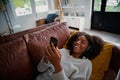 Woman with toothy smile lying on couch using smartphone at home listening to music on bluetooth wireless headphones Royalty Free Stock Photo