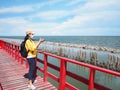 Woman tourist wearing face mask standing on red bridge and taking photograph summer beach