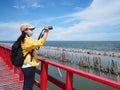 Woman tourist wearing face mask standing on red bridge and taking photograph summer beach