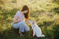 Woman training cute white puppy to behave and caressing him in summer meadow in warm sunset light. Adorable fluffy puppy looking