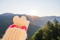 Woman traveler in christmas gloves holding snow in the shape of a heart on a background of mountains with sunbeams. Winter travell