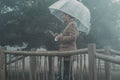 Woman use mobile phone under the rain protected from a transparent umbrella. Rainy day and bad weather condition at the park.