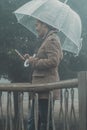 Woman use mobile phone under the rain protected from a transparent umbrella. Rainy day and bad weather condition at the park.