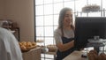 Woman using computer in bakery with pastries on display and happy customer present Royalty Free Stock Photo