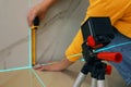 Woman using cross line laser level and tape for accurate measurement on tiled wall, closeup