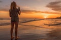 Woman using her mobile phone while taking photos of the sunset on the beach. Royalty Free Stock Photo